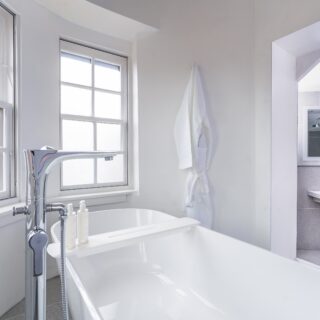 A modern hotel room features a large bed with white linens, a wooden headboard, nightstands, a wall-mounted television, and a window with city views.