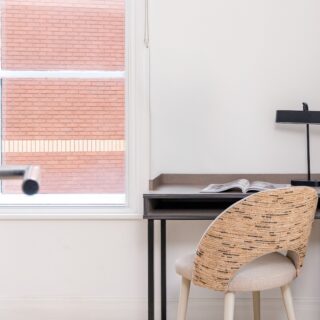 Street view of a modern hotel building with large windows, a prominent red sign, and trees in the foreground, located in an urban area with bright sunshine.