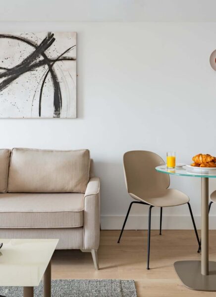 The image shows a modern, elegantly furnished hotel suite with a large bed, stylish chairs, a desk, and large windows providing a cityscape view.