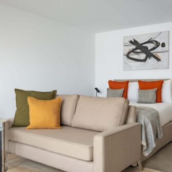 The image features a stylish modern studio apartment with minimalistic decor, including a sleek sofa, contemporary floor lamp, wall art, and large windows with sheer curtains.
