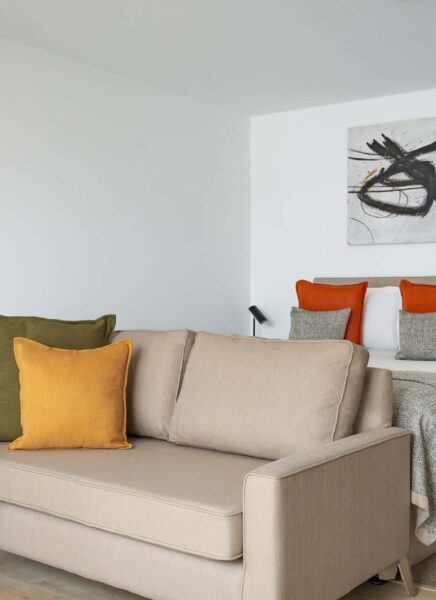 The image features a stylish modern studio apartment with minimalistic decor, including a sleek sofa, contemporary floor lamp, wall art, and large windows with sheer curtains.