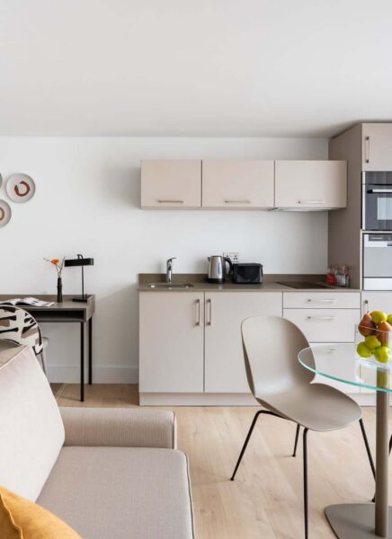 A cozy, apartment interior featuring a living area with a grey sofa, decorative cushions, and a modern circular coffee table by a floor-to-ceiling window draped with white curtains.