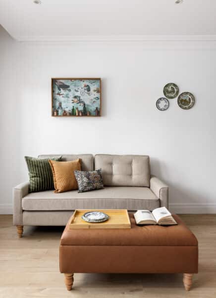 A modern studio apartment featuring a cozy bed, wooden flooring, minimalist décor, wall-mounted art, a sitting area with a table and chairs, and large windows for natural light.