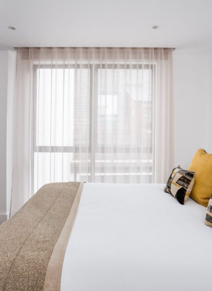 Modern living room with large windows, comfy grey sofas, a small round table, and a decorative plant. Warm lighting and wooden flooring create a cozy and inviting atmosphere.