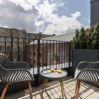 A cozy, modern studio apartment with a neatly made bed, kitchenette, small dining table, framed artwork, and a large window illuminating the space with natural light.