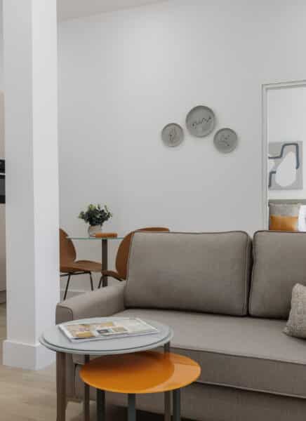 A modern, stylish living room with a sleek gray sofa, glass coffee table, and abstract artwork on the wall. Large windows let in natural light, overlooking greenery.