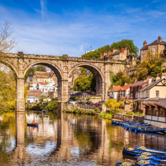 The image shows a scenic view of Knaresborough, with a picturesque viaduct reflected in the river below, surrounded by lush greenery and historic buildings under a clear blue sky.