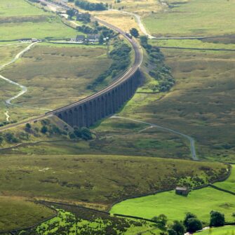 A historic stone arch railway viaduct stretches across a lush green valley under a bright blue sky with scattered white clouds.