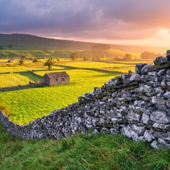 The image shows a scenic view of the Yorkshire Dales with rolling green hills, scattered trees, stone walls, and a cloudy sky.