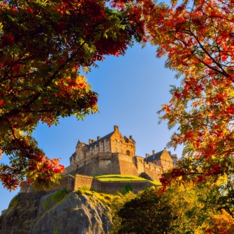 A historic cityscape of Edinburgh featuring the iconic Edinburgh Castle atop Castle Rock with surrounding buildings under a partly cloudy sky.