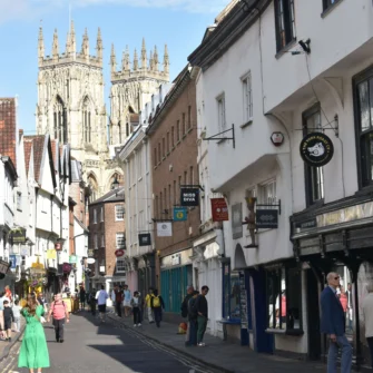 Street view of a bustling shopping district in York, featuring historic architecture, numerous pedestrians, and storefronts, with a clear sky in the background.