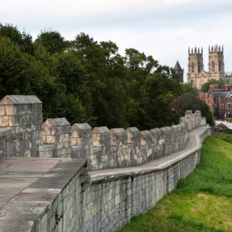 A historic stone city wall stretches into the distance, surrounded by lush greenery and tree-lined paths, under a clear blue sky.